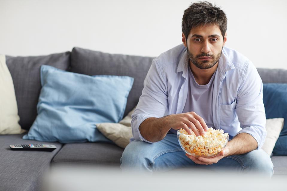 Portrait of sad bearded man watching TV and holding bowl of popcorn while sitting on sofa at home